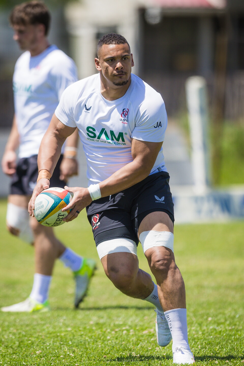 A rugby player in a white shirt and black shorts runs with a ball.
