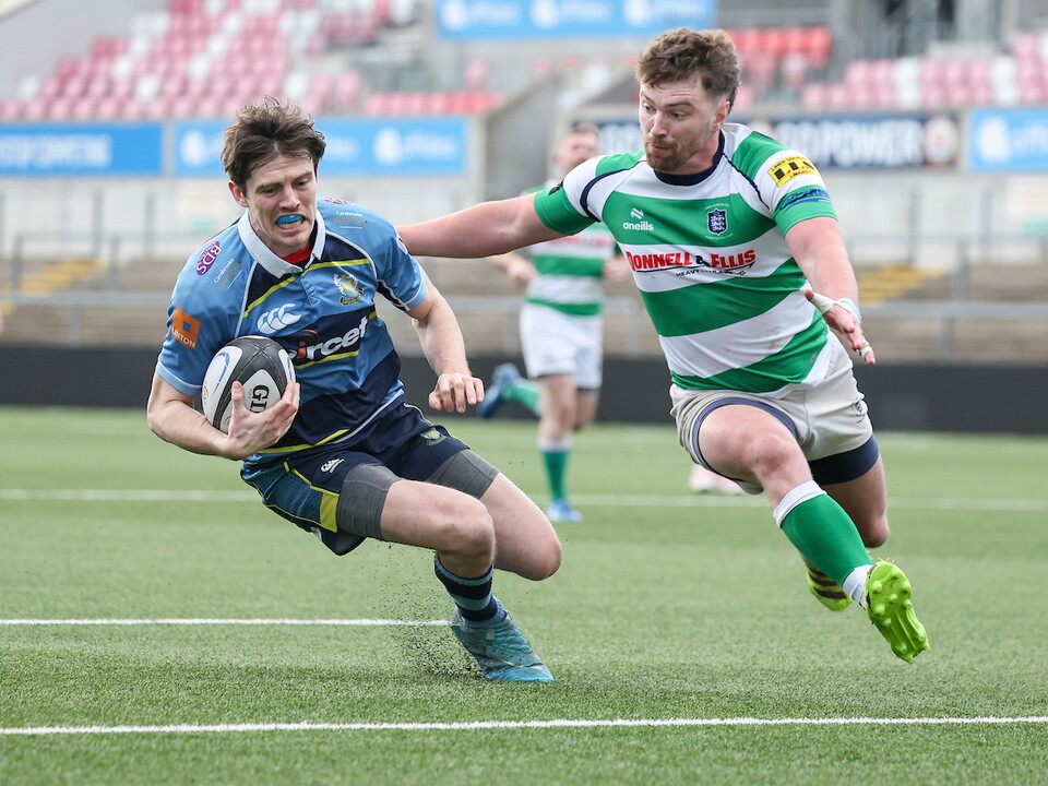 Two players in blue and green striped kits run across a rugby pitch, one holding a ball.